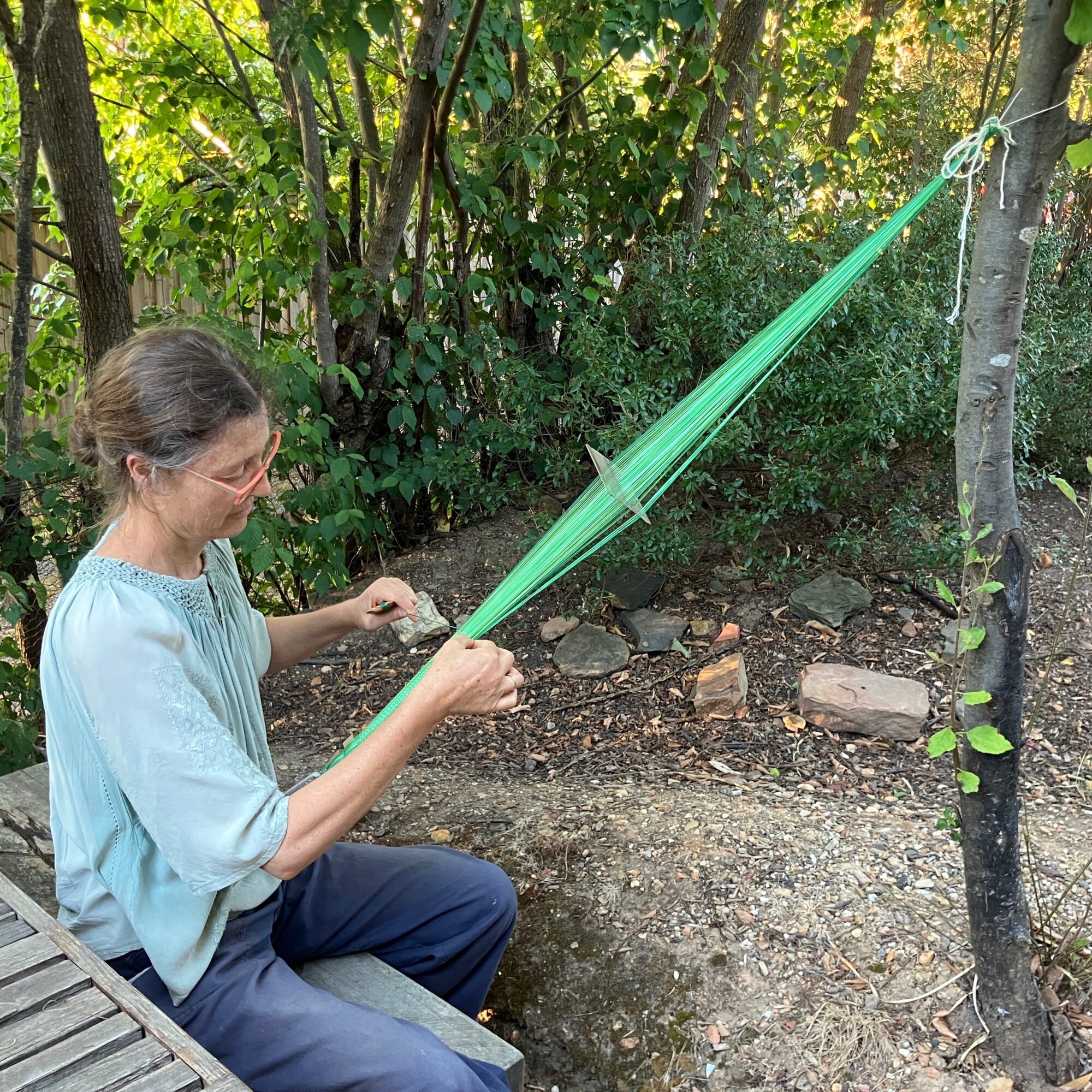 A slender white woman, Ilka White, is weaving a narrow green band. The end of her weaving is attached to a tree and there are shrugs in the background