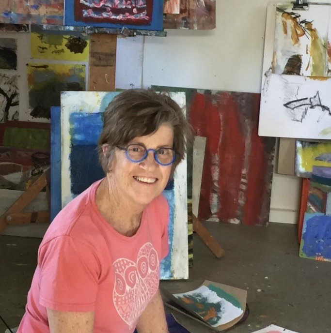 Prue McAdam, at artist, is looking at the camera, smling. She is wearing a coral coloured T-shirt and artworks are behind her.